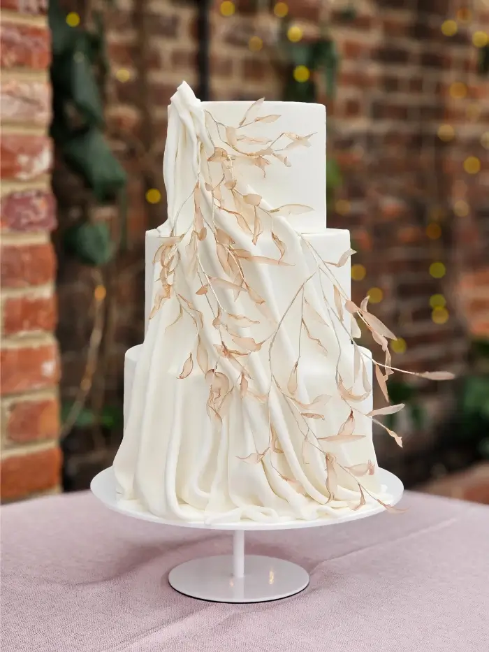Contemporary 3 tier autumal wedding cake. White textured tiers with silky drape from top to bottom and glassy effect golden leaves for this Northbrook Park wedding.